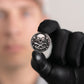 A person in black gloves holds up The Omen Classic Antique, Diamond & Silver Edition coin by Doubloon, featuring a skull and crossed swords with "Stackin’ the Silver" inscribed; their face is blurred in the background.