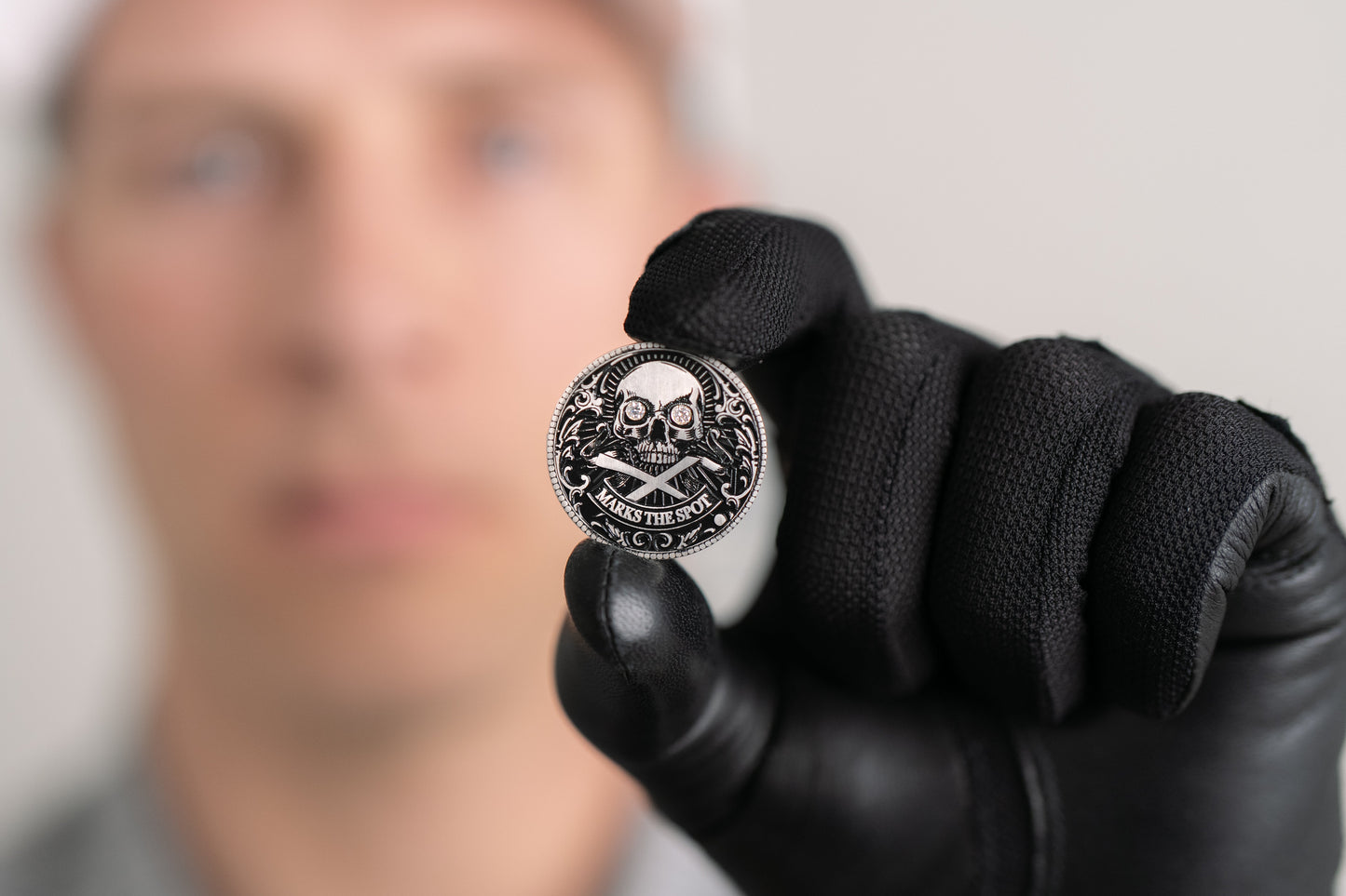 A person in black gloves holds up The Omen Classic Antique, Diamond & Silver Edition coin by Doubloon, featuring a skull and crossed swords with "Stackin’ the Silver" inscribed; their face is blurred in the background.