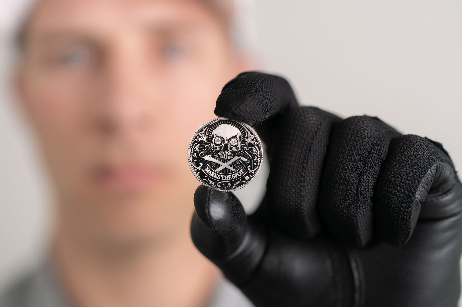 A person in black gloves holds up The Omen Classic Antique, Diamond & Silver Edition coin by Doubloon, featuring a skull and crossed swords with "Stackin’ the Silver" inscribed; their face is blurred in the background.