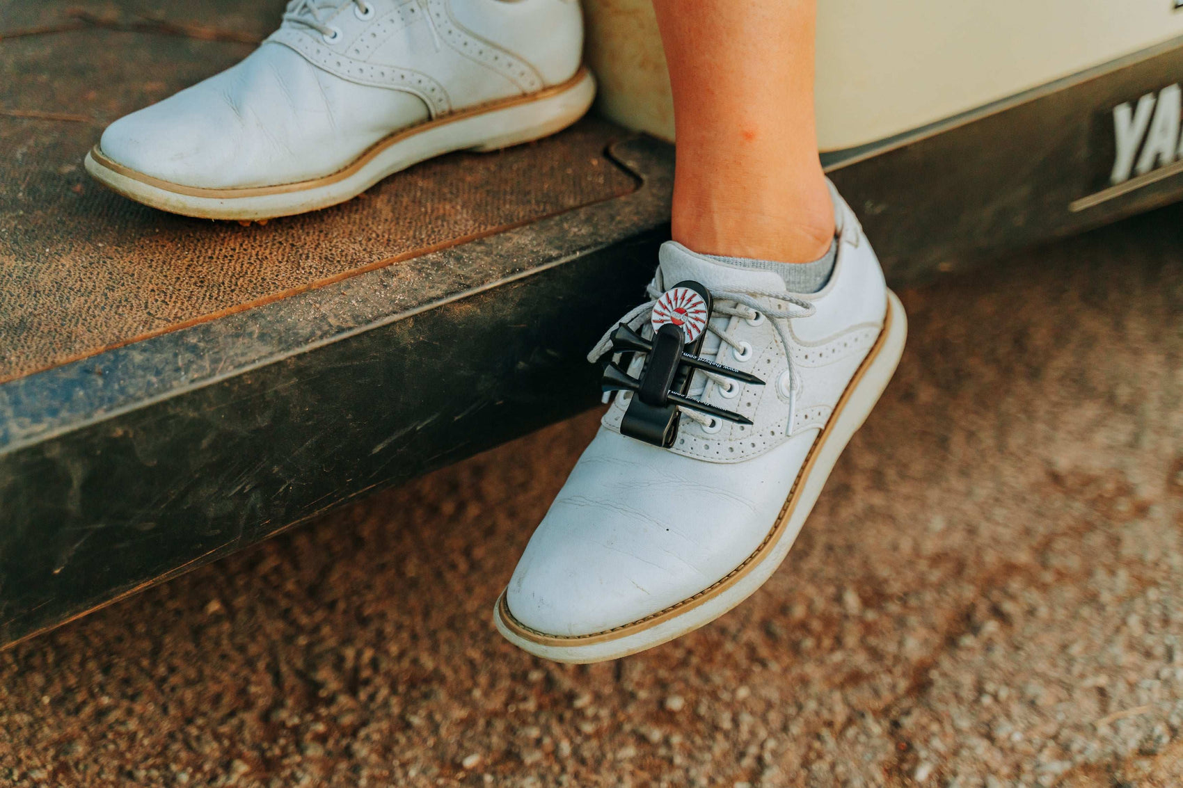 A close-up of a person’s foot wearing THE BYRD GOLF CLYP - Red Cardinal by The Byrd, LLC, a stylish golf accessory attached to the laces of a light blue saddle shoe resting on the edge of a vehicle or golf cart.