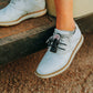 Close-up of a person in light blue saddle shoes with white soles, sitting on an edge. Their laces are fastened with THE BYRD GOLF CLYP - Black Byrd by The Byrd, LLC, which displays a small circular decorative emblem.