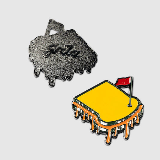 Two enamel Pimento Ball Markers by Guerrilla Golf, shaped like melting grilled cheese sandwiches: one features a yellow design with an orange cheese drip and a small red flag, while the other displays a black metal back engraved with "grls," resembling handwriting from an edible marker. Both are showcased against a gray background.