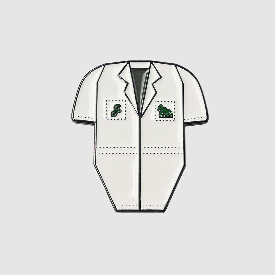 A Guerrilla Golf Caddy Bib Ball Marker pin shaped like a white short-sleeve lab coat, featuring green symbols on the chest pockets, reminiscent of a fun novelty marker and set against a plain gray background.