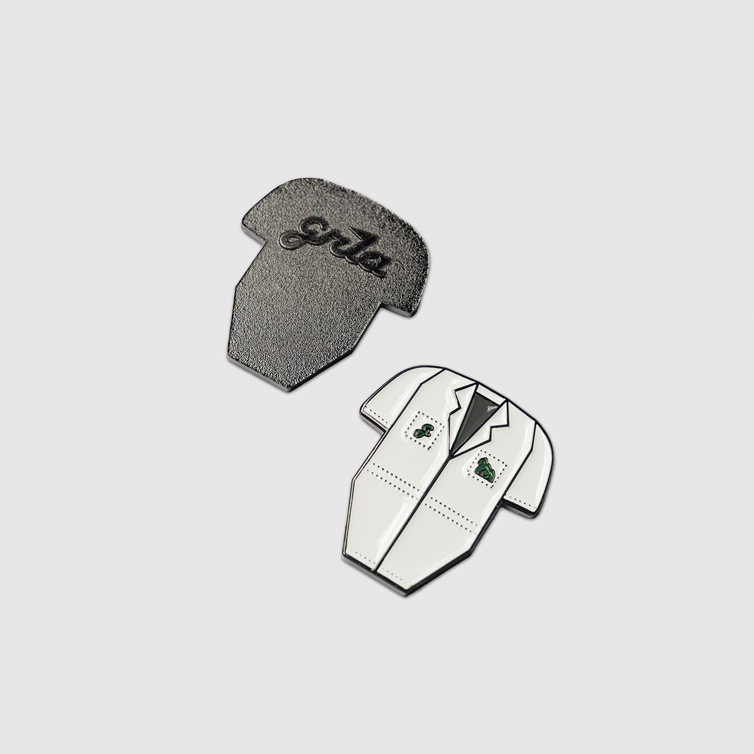Two enamel pins—one shaped like a gray shirt with "Girls" written on it, the other shaped like a white suit jacket with black lapels and green detailing—both designed by Guerrilla Golf using food-safe marker for added precision, from the Caddy Bib Ball Marker collection, are displayed on a plain white background.