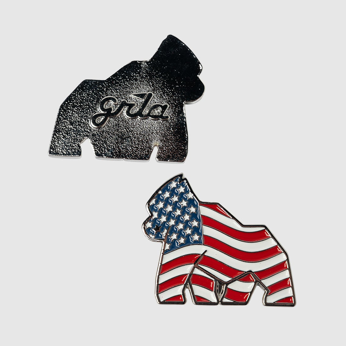 Two gorilla-shaped ball markers from Guerrilla Golf are displayed on a light gray background, evoking the feel of classic golf memorabilia: one, the American Flag Gorilla Ball Marker, showcases a bold stars and stripes design; the other is plain silver with "gorilla" engraved on it.