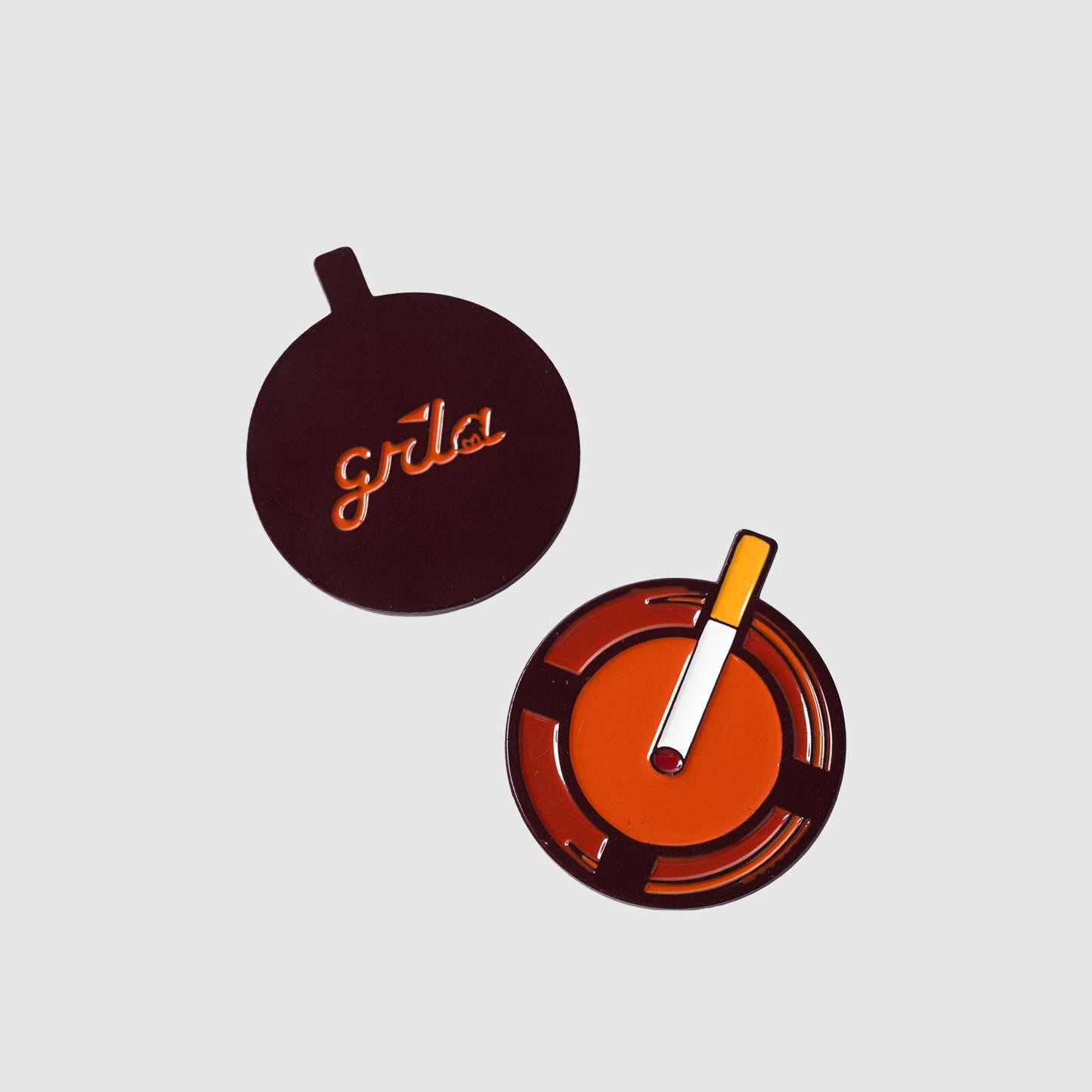 Two round objects on a light background: one is the Guerrilla Golf Retro Ashtray Ball Marker with a cigarette detail, and the other is a matching orange and dark brown lid featuring "grla" branding. This quirky set makes a funny golf gift for anyone who loves unique accessories.