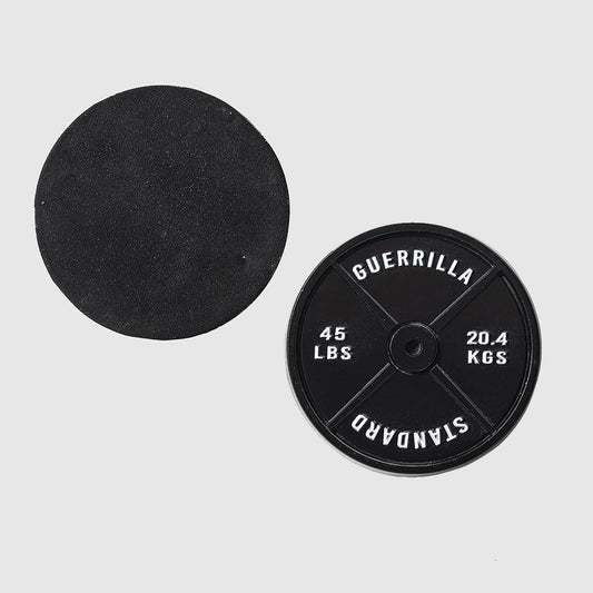 Two black Guerrilla Golf 45lb Plate Ball Markers on a light gray background; one marker displays the GUERRILLA STANDARD 45 LBS 20.4 KGS text, while the other is turned over, its blank side resembling a manhole cover.