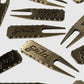A set of Guerrilla Golf GRLA Raw Divot Tools—bronze-colored metal golf accessories with scalloped edges and the GRLA logo engraved on each—arranged stylishly against a plain light background.