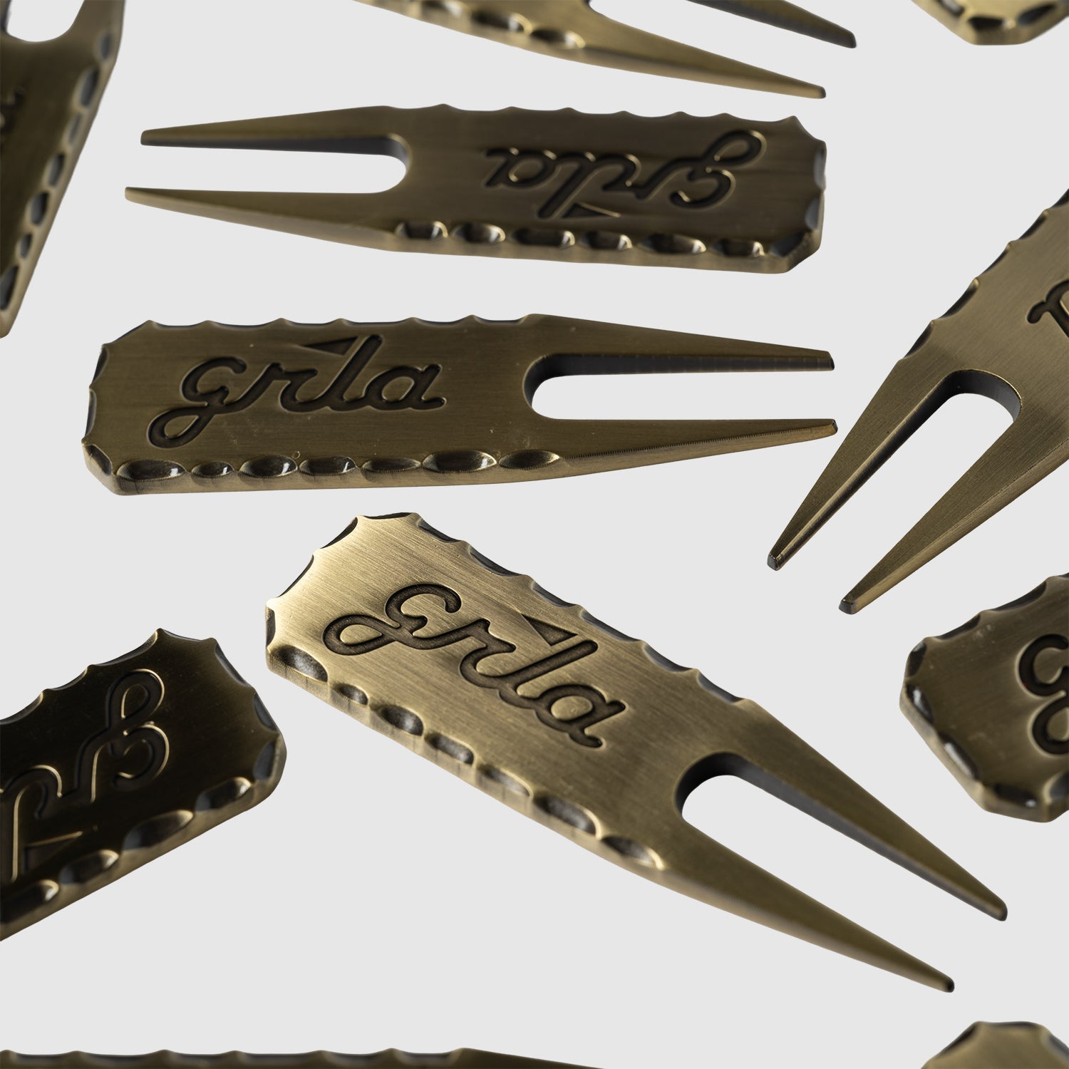 A set of Guerrilla Golf GRLA Raw Divot Tools—bronze-colored metal golf accessories with scalloped edges and the GRLA logo engraved on each—arranged stylishly against a plain light background.