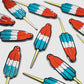 Guerrilla Golf’s Rocket Pop Divot Tool, featuring vibrant red, white, and blue rocket-shaped designs with yellow pin backs, offers a playful nod to childhood nostalgia—making it the perfect golf accessory for anyone looking to add a whimsical touch to their game.