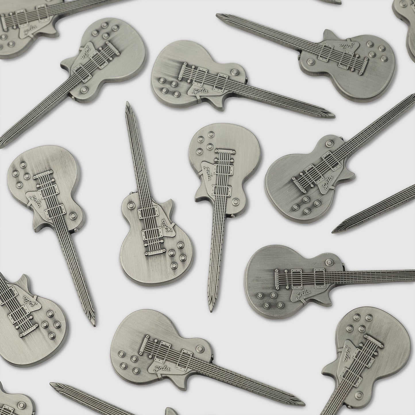 Several Guerrilla Golf Guitar Divot Tools, each crafted from metal and shaped like an electric guitar with embossed details such as strings, knobs, and pickups, are scattered on a white surface—making them the perfect accessory for golf enthusiasts who want to add a musical twist to their game.