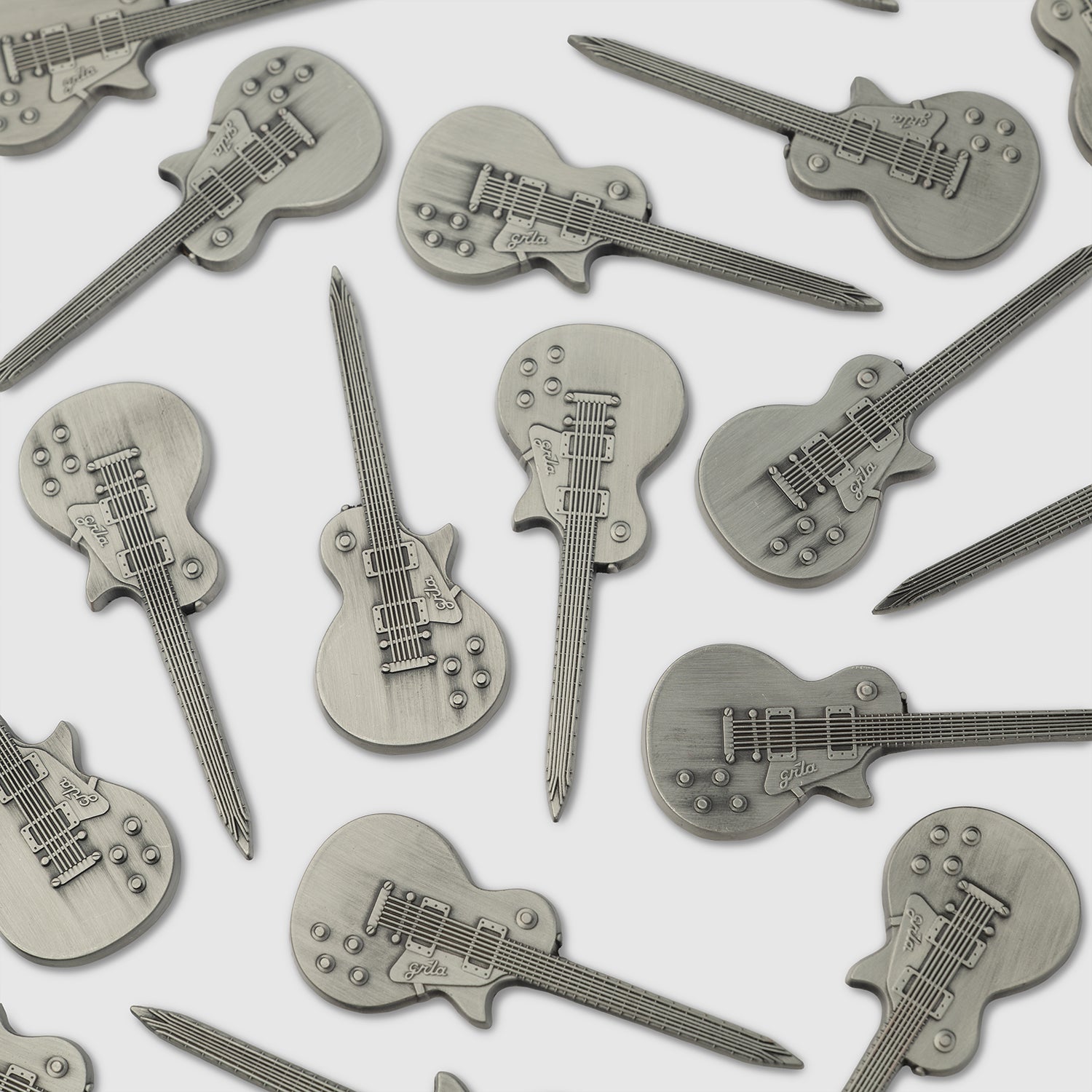 Several Guerrilla Golf Guitar Divot Tools, each crafted from metal and shaped like an electric guitar with embossed details such as strings, knobs, and pickups, are scattered on a white surface—making them the perfect accessory for golf enthusiasts who want to add a musical twist to their game.