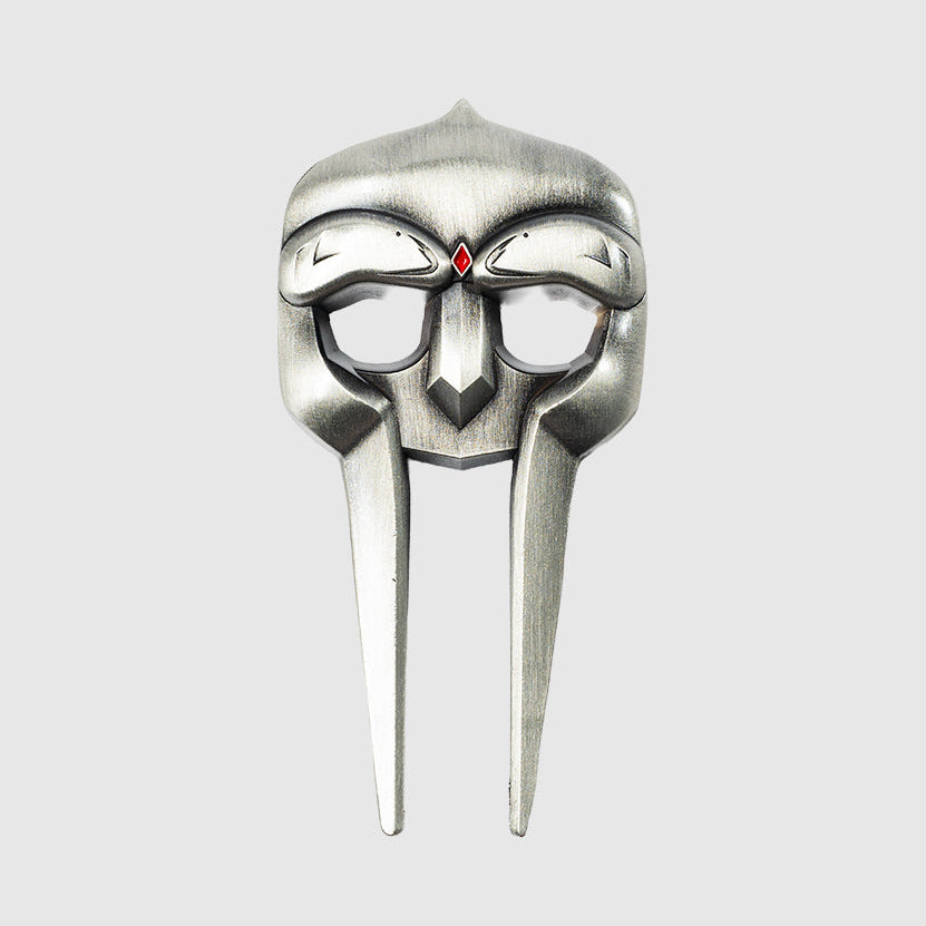 The Gladiator Divot Tool by Guerrilla Golf features a metallic mask design with a striking silver finish, two eye holes, a central red gem above the nose area, and two elongated vertical sections extending downward—resembling a sleek golf accessory for repairing divots on the green—prominently displayed against a plain gray background.