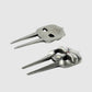 The Gladiator Divot Tool by Guerrilla Golf features two distinct metal designs, each with two prongs: one showcases a flat, skull-like shape with intricate cutouts and engraved text, while the other boasts a more sculpted, detailed skull motif. Both uniquely crafted golf accessories are designed to repair divots on the course and are displayed resting on a plain, light gray surface.