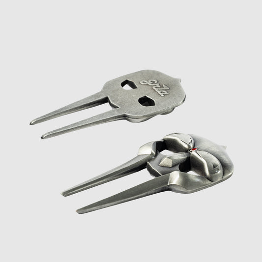 The Gladiator Divot Tool by Guerrilla Golf features two distinct metal designs, each with two prongs: one showcases a flat, skull-like shape with intricate cutouts and engraved text, while the other boasts a more sculpted, detailed skull motif. Both uniquely crafted golf accessories are designed to repair divots on the course and are displayed resting on a plain, light gray surface.