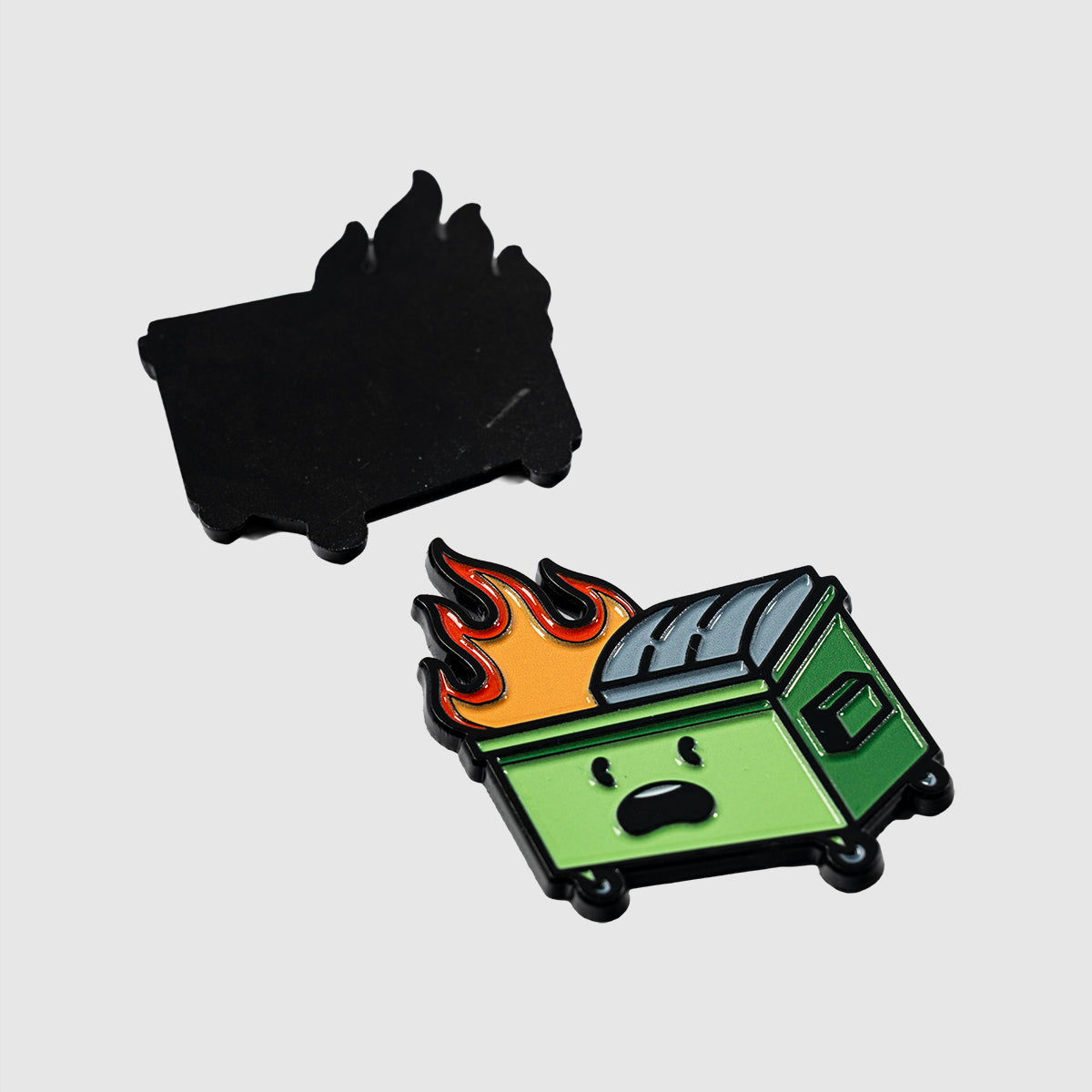 The Dumpster Fire enamel pin by Guerrilla Golf features a playful green dumpster ablaze, complete with a surprised expression and finished in shiny black nickel. Its vibrant enamel colors and flame-shaped solid black backing make it a whimsical twist on the classic dumpster fire motif.