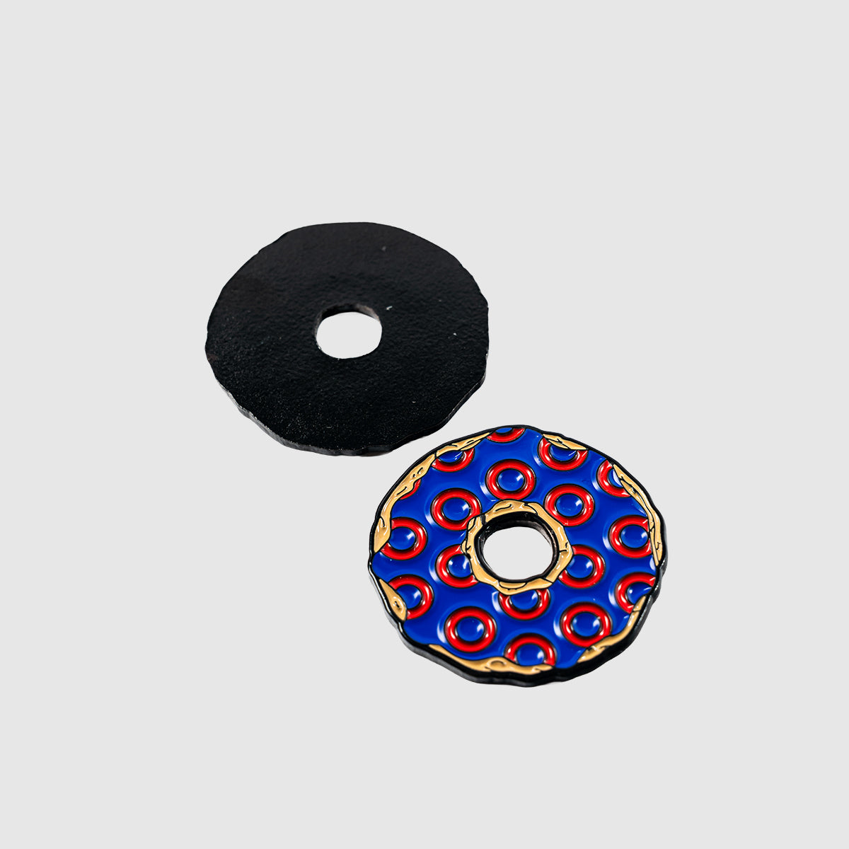 Two Donut Ball Markers by Guerrilla Golf are on display: one features a solid black design, while the other is adorned with a blue and red pattern accented with gold circles. Both markers are presented against a plain, light background.