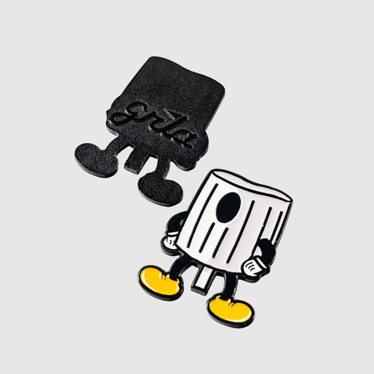 Two cartoonish enamel pins from Guerrilla Golf, called the Fat Cap Ball Marker, draw inspiration from graffiti and NY fat styles. Shaped like trash cans with arms, legs, and yellow shoes, one design is black and says "girls," while the other is white with black details and features a single large eye on the can. These quirky accessories double as playful ball markers, shown against a plain gray background.