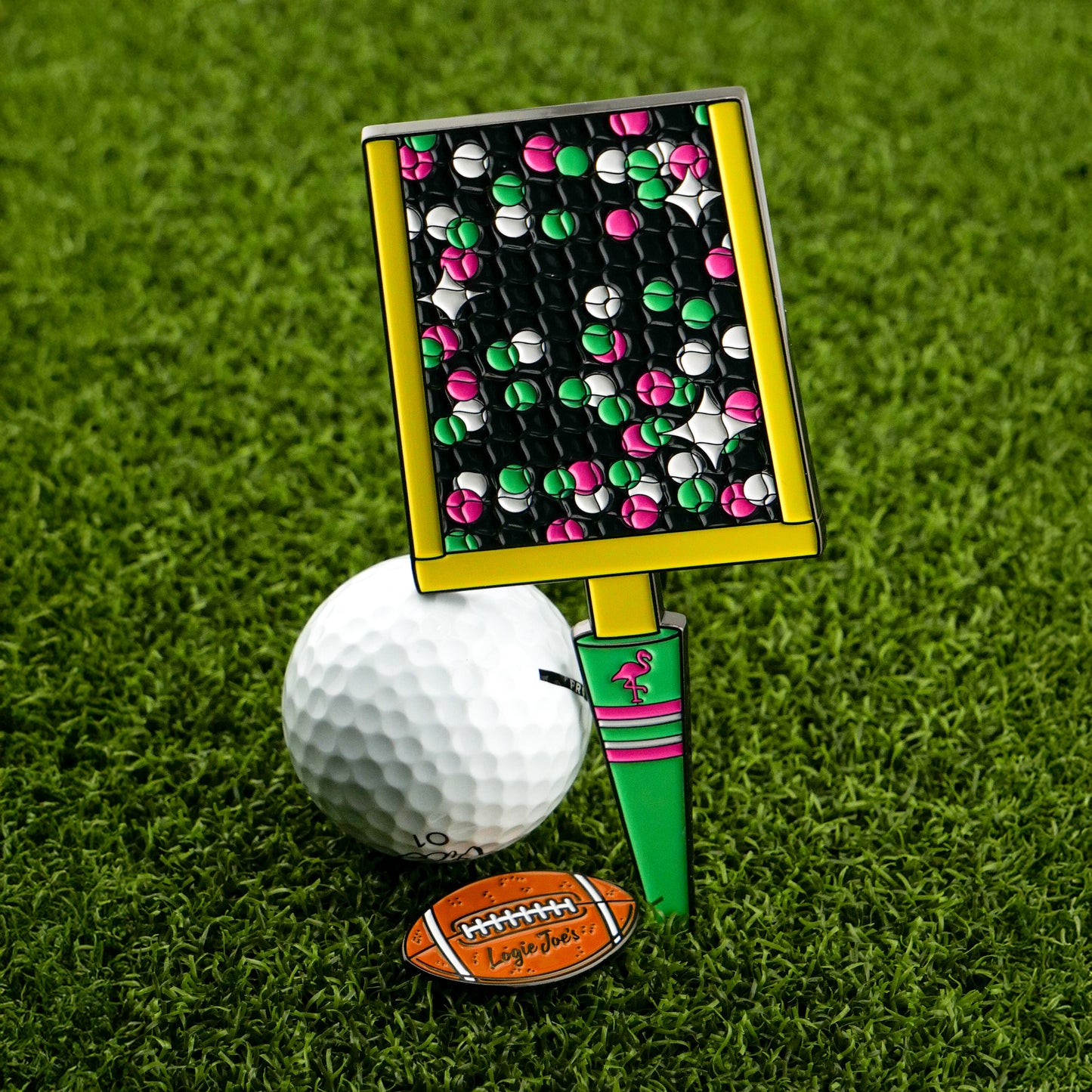 A Logie Joe's Golf Field Goal Divot Tool with a colorful, rectangular design includes a removable football ball marker, resting on grass.