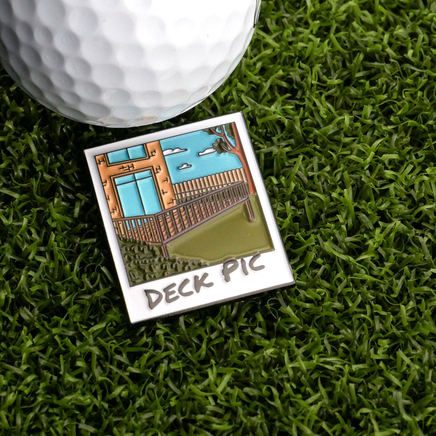 The Deck Pic Ball Marker by Ballmarkers.com, featuring a cartoon backyard deck and blue sky, rests on green artificial grass next to a golf ball.