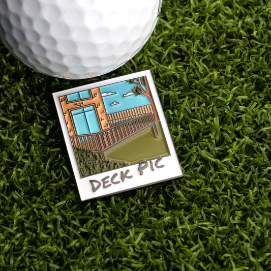 The Deck Pic Ball Marker by Ballmarkers.com, featuring a cartoon backyard deck and blue sky, rests on green artificial grass next to a golf ball.