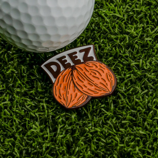 A white golf ball rests on green turf beside Logie Joe's Golf Deez Nuts Ball Marker, an enamel pin with two brown walnuts and 