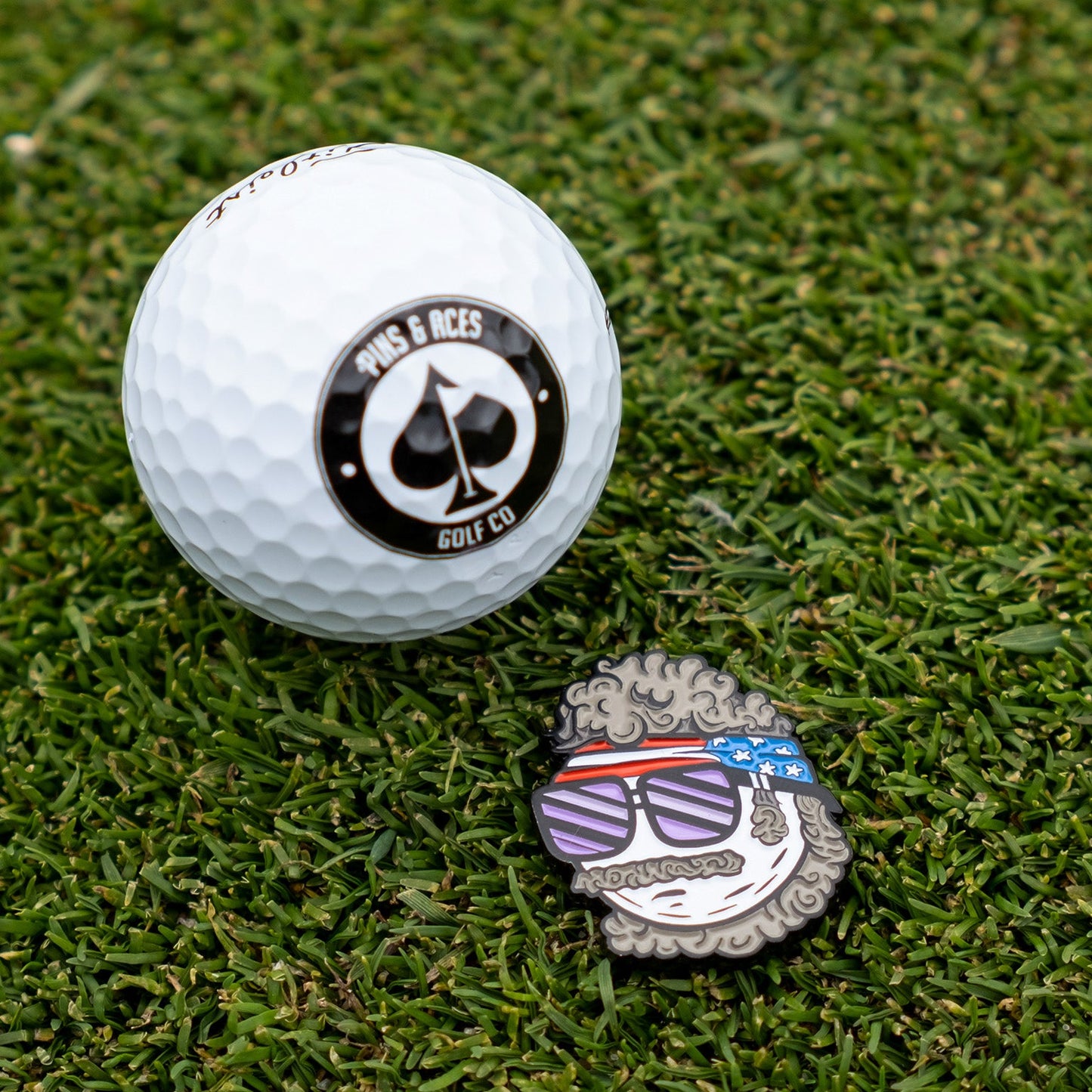 A Pins and Aces golf ball rests on grass beside the Jerry the Degenerate Ball Marker, a novelty enamel marker featuring a cartoon face with curly hair, sunglasses, and an American flag headband.