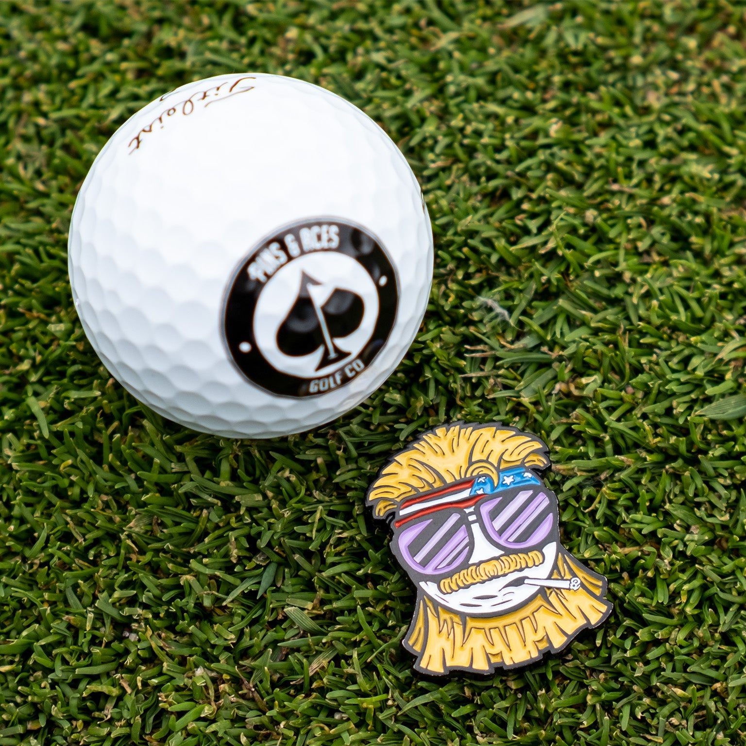 The Dale the Degenerate Ball Marker by Pins and Aces features a colorful cartoon bearded man pin with sunglasses, headband, and cigarette, next to a white golf ball with a spade logo—perfect as fun magnetic ball markers on the green.