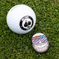 A Pins and Aces golf ball and the Gus the Degenerate Ball Marker, shaped like a bearded cartoon with sunglasses and a headband, rest on grass. The golf ball displays a black and white Birds & Aces Golf Co. logo.