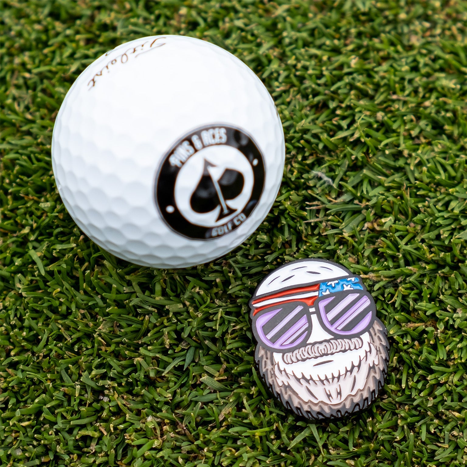 A Pins and Aces golf ball and the Gus the Degenerate Ball Marker, shaped like a bearded cartoon with sunglasses and a headband, rest on grass. The golf ball displays a black and white Birds & Aces Golf Co. logo.
