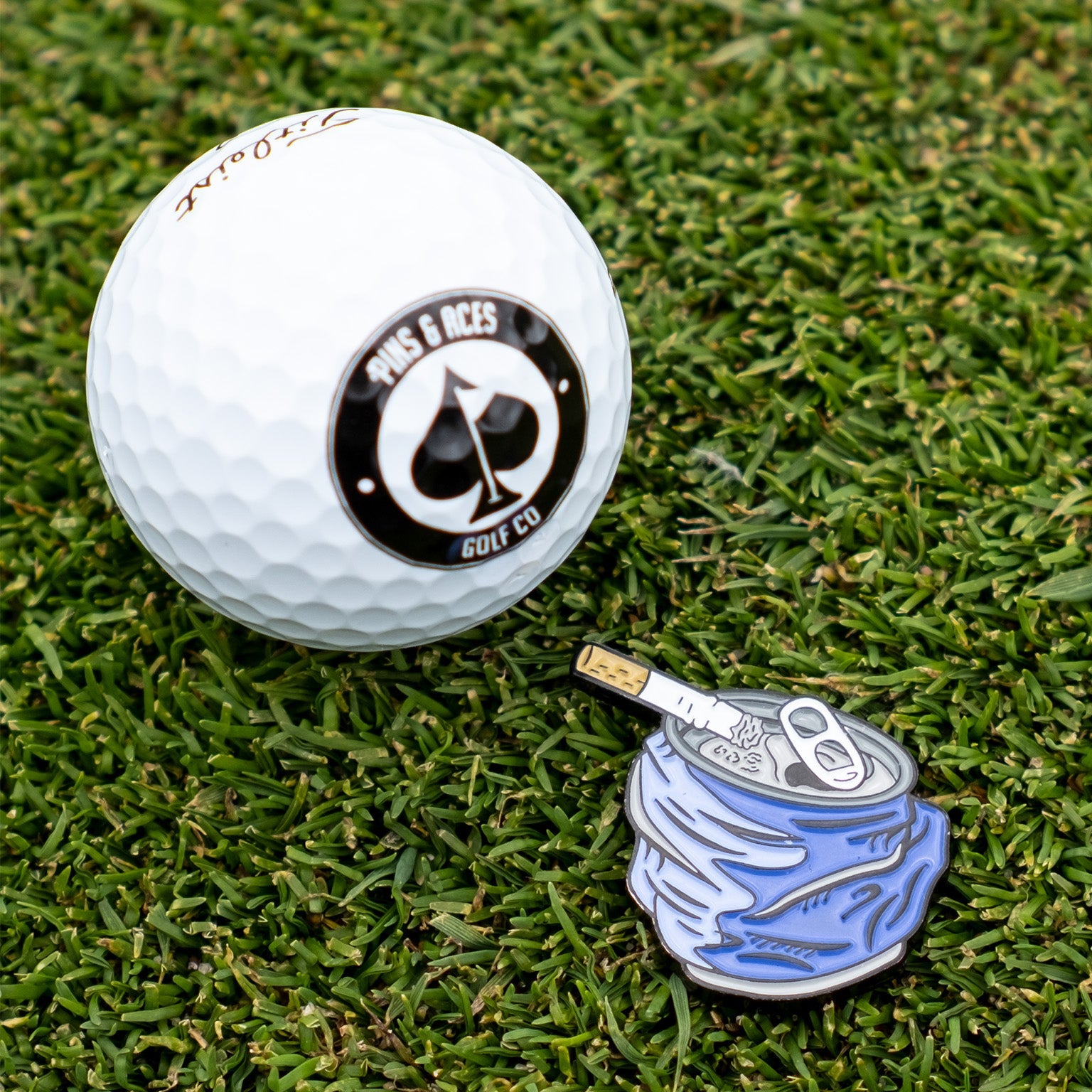 A white golf ball with a black and white 7als & Aces Golf Co. logo sits beside the Pins and Aces Crushed Can Ball Marker with enamel paint, both resting on green grass.