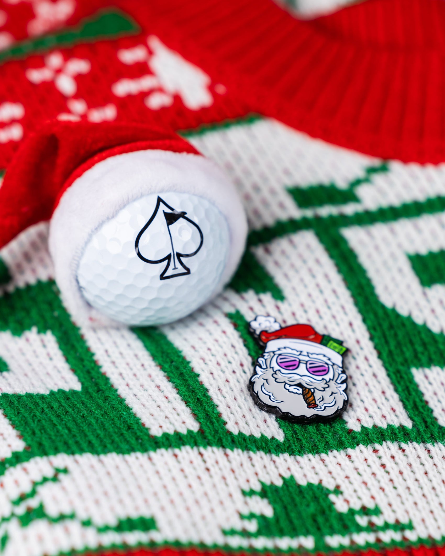 The Pins and Aces "Santa the Degenerate Ball Marker" features a magnetic Santa golf ball marker, Santa hat, and sunglasses-wearing Santa pin—ideal Christmas golf accessories on a festive red, white, and green sweater.