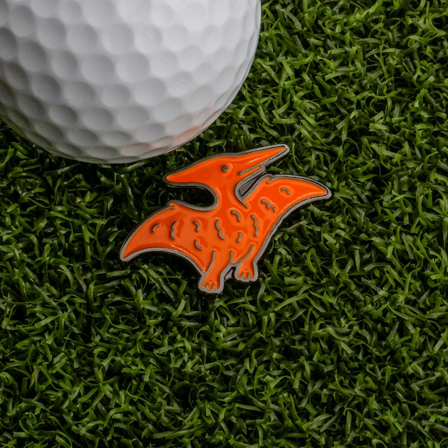A white golf ball and the Ballmarkers.com Pterodactyl Dinosaur Nugget Ball Marker sit on green artificial grass, bringing playful charm to your golf accessories.