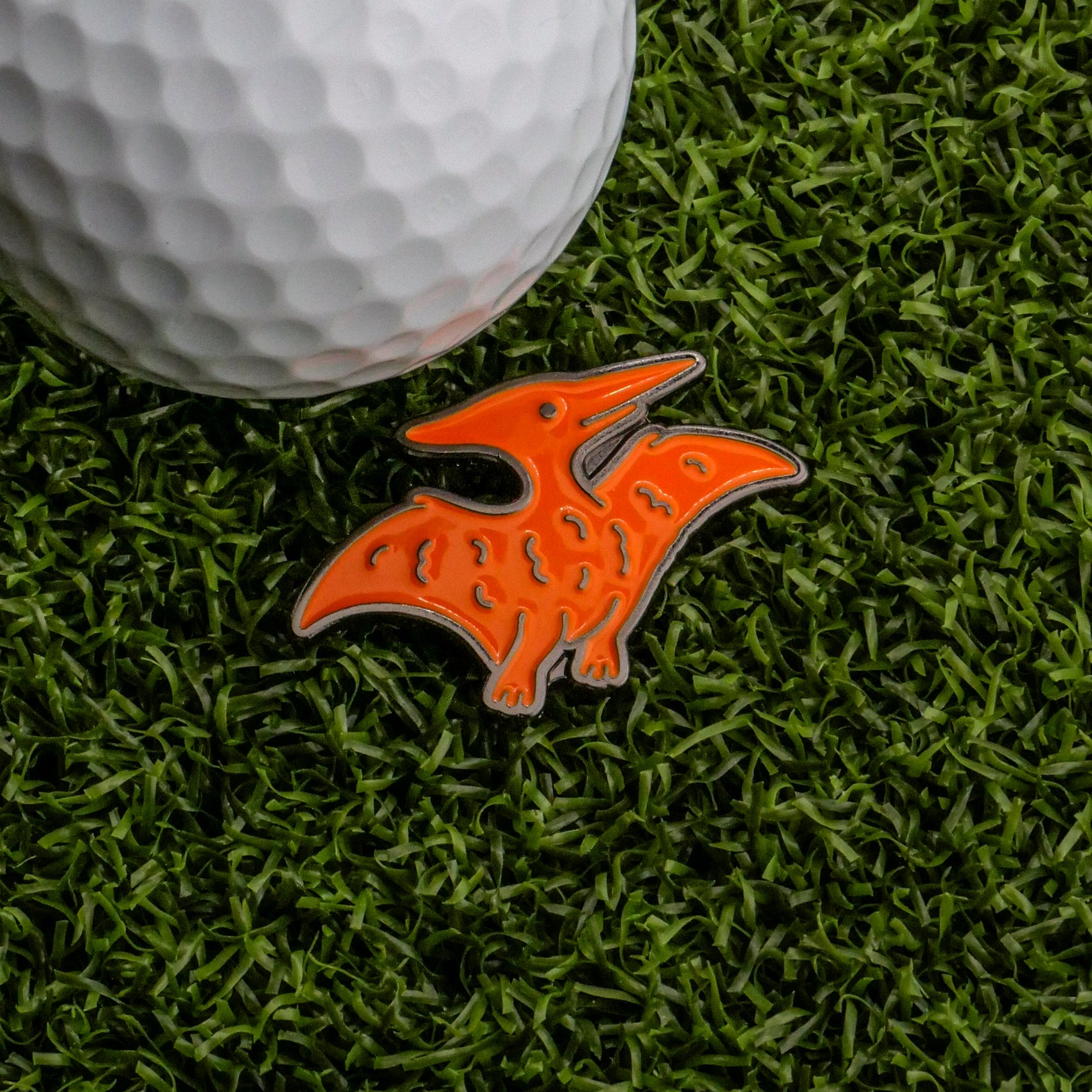 A white golf ball and the Ballmarkers.com Pterodactyl Dinosaur Nugget Ball Marker sit on green artificial grass, bringing playful charm to your golf accessories.