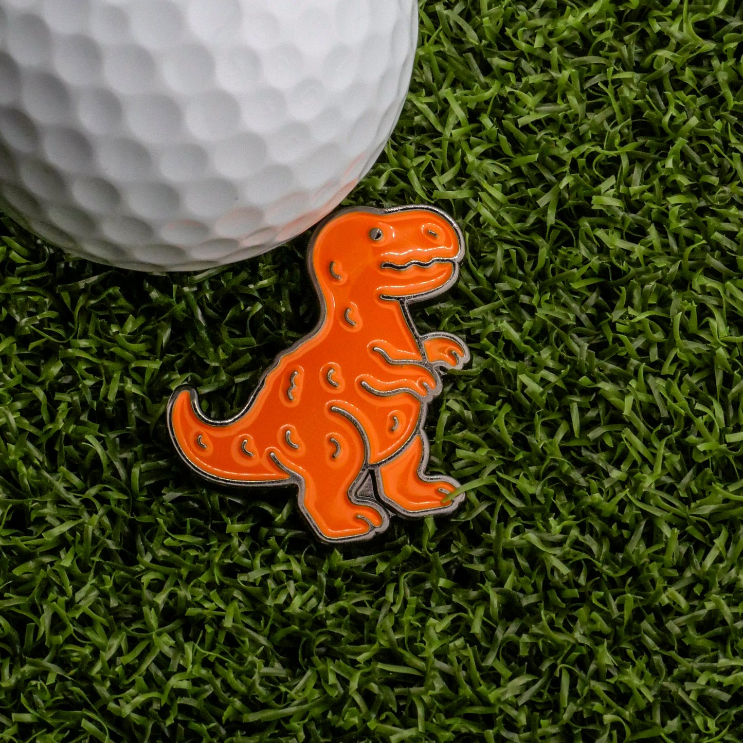 A close-up of a white golf ball on artificial green grass beside the Ballmarkers.com T-Rex Dinosaur Nugget Ball Marker, which features a smiling orange T-Rex.