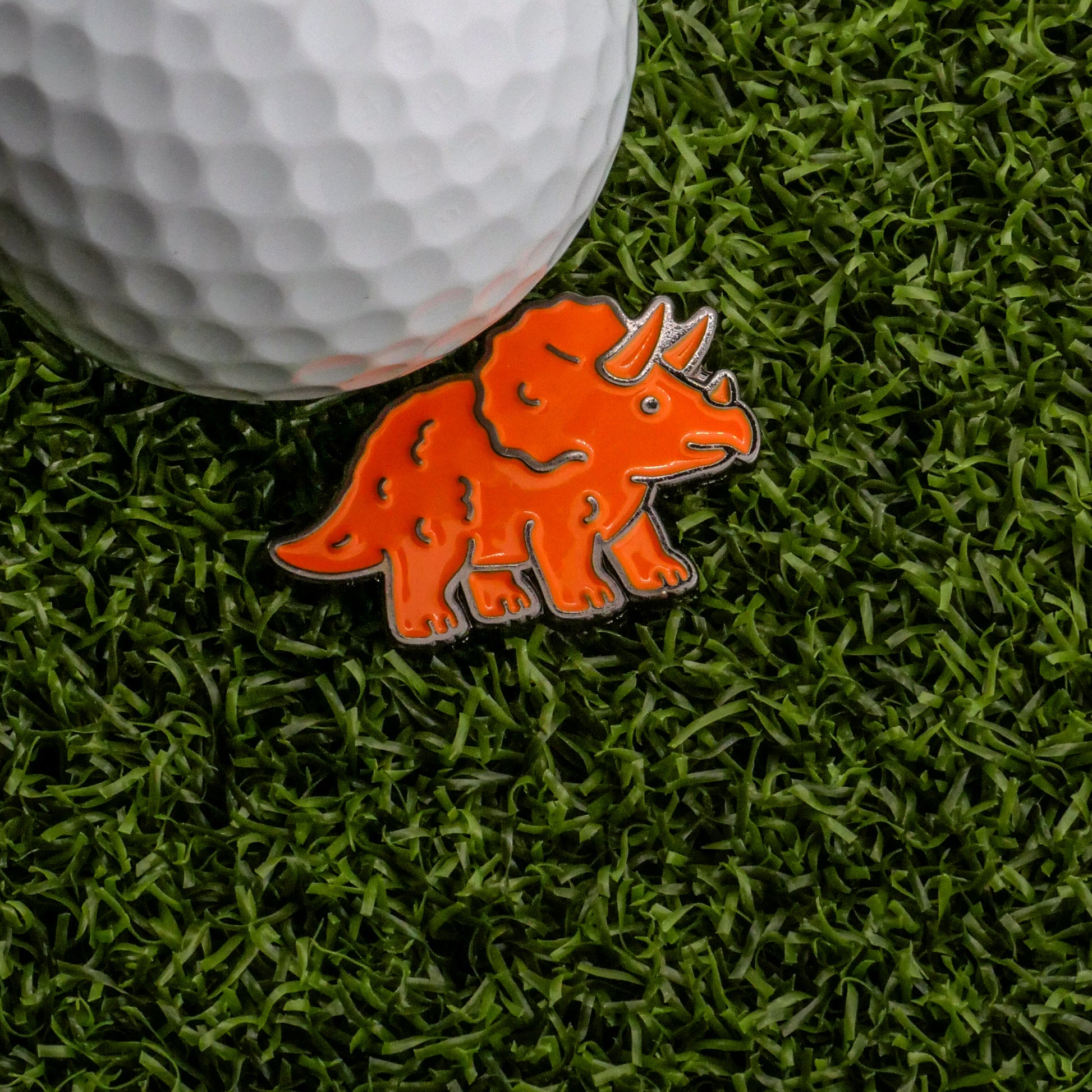 A white golf ball on artificial grass beside the Triceratops Dinosaur Nugget Ball Marker by Ballmarkers.com—the perfect golf accessory for dino nuggies fans.