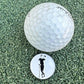 A Grunk Dolfer Grunk Divot Tool, a white golf ball, and a round ball marker with a golfer silhouette are displayed on artificial green turf.