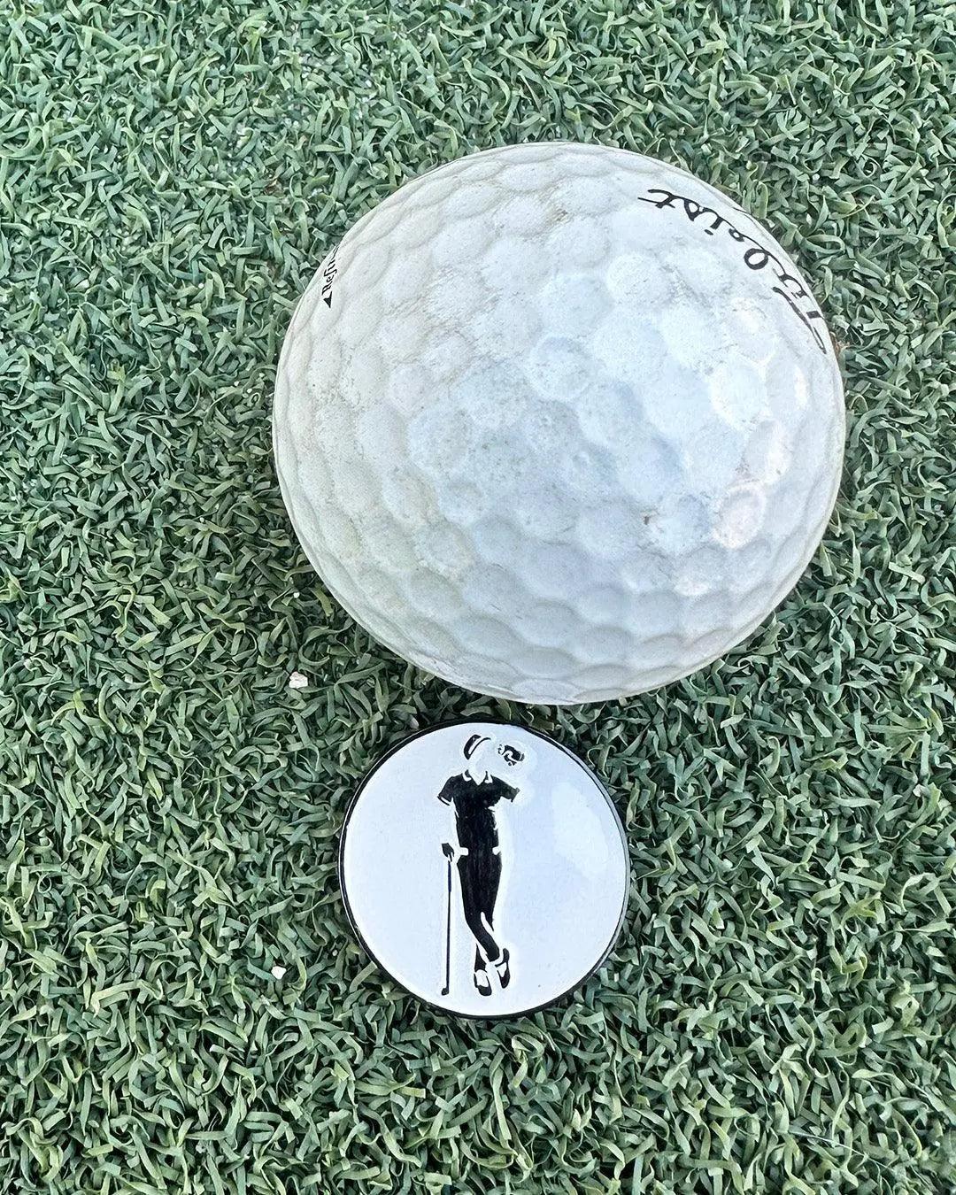 A Grunk Dolfer Grunk Divot Tool, a white golf ball, and a round ball marker with a golfer silhouette are displayed on artificial green turf.