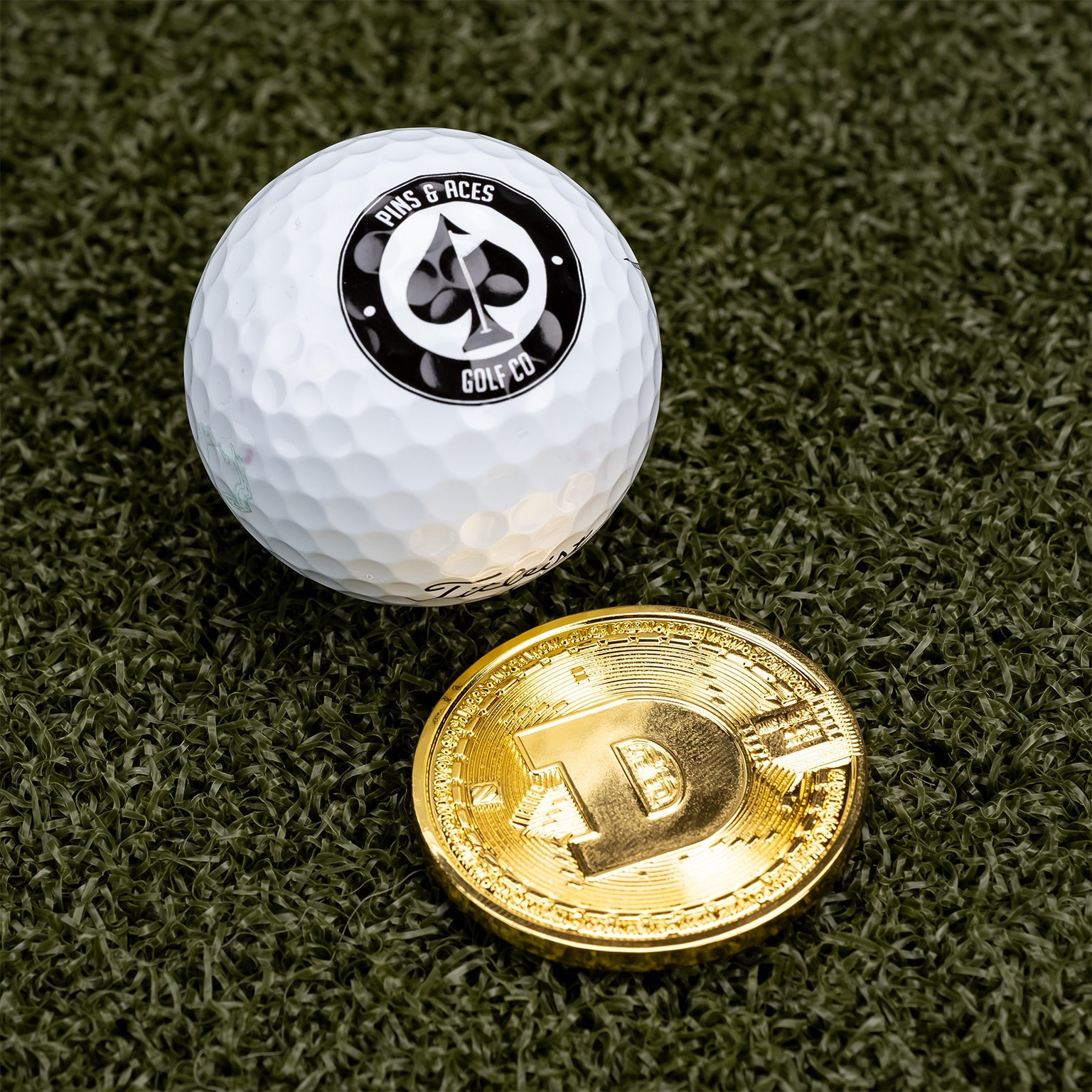 A white golf ball with a black Pins and Aces logo rests beside the Dogecoin Ball Marker—a shiny, gold chrome token—on green artificial grass.