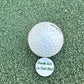 The Grunk Dolfer Donde Esta Ball Marker sits on white artificial turf next to a golf ball, featuring “Donde Esta La Cart Girl” in green lettering—a fun golf accessory for your game.