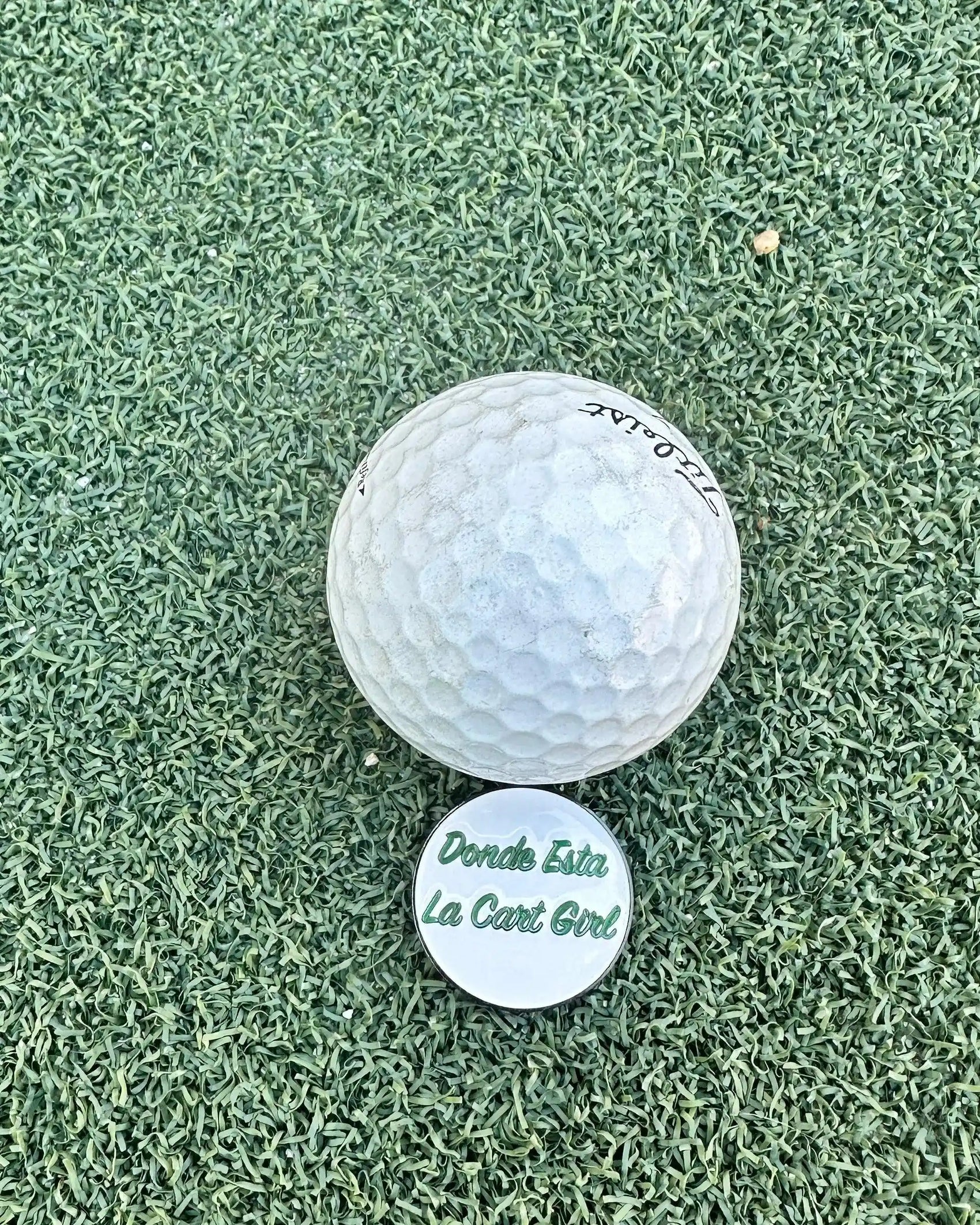 The Grunk Dolfer Donde Esta Ball Marker sits on white artificial turf next to a golf ball, featuring “Donde Esta La Cart Girl” in green lettering—a fun golf accessory for your game.