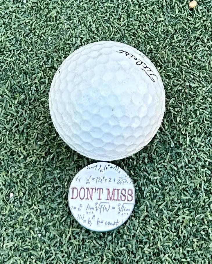 The Grunk Dolfer Don't Miss Ball Marker, featuring bold red text and surrounded by handwritten math formulas, sits on artificial grass beside a golf ball—making it a perfect unique golf gift.