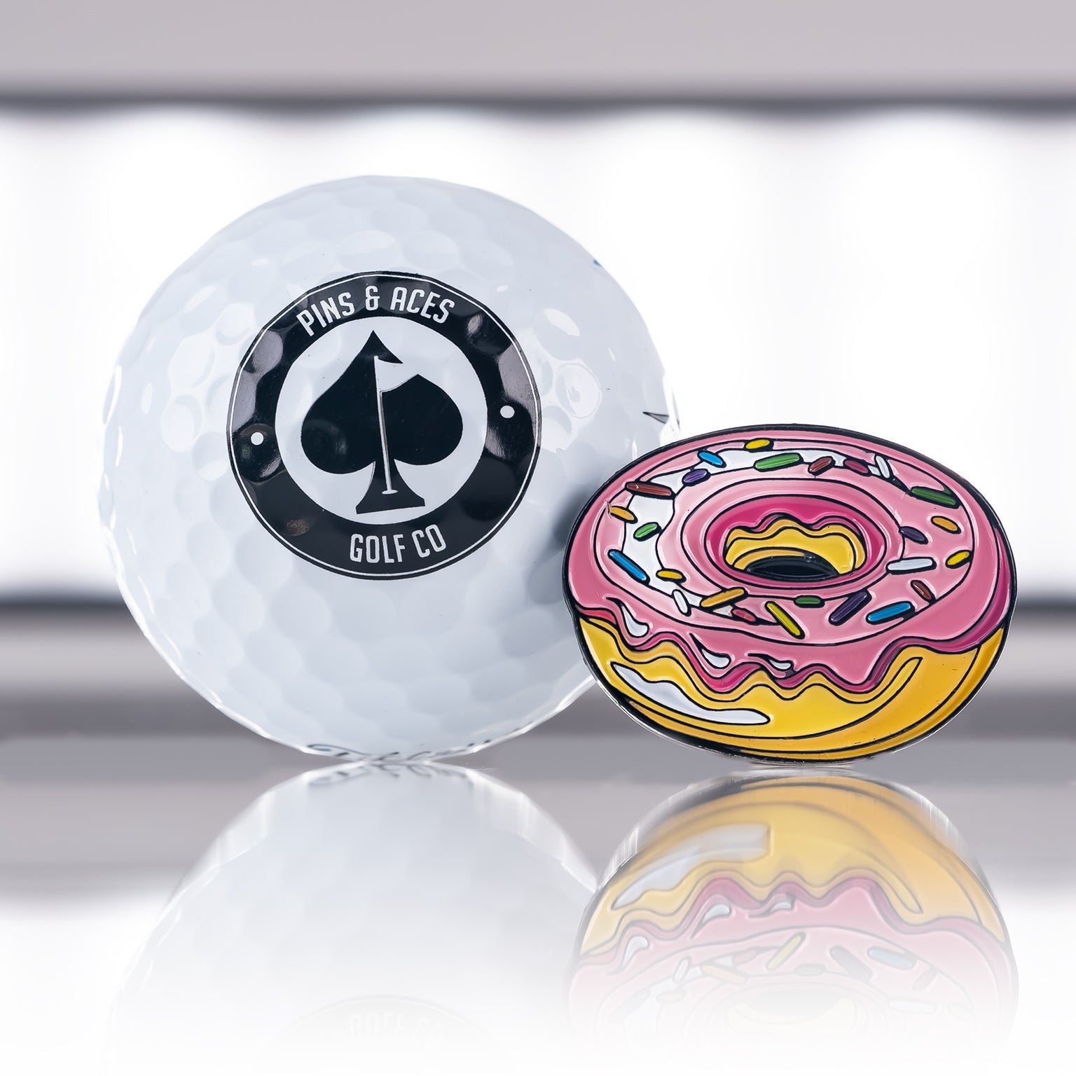 A white Pins and Aces golf ball sits beside the Donut Ball Marker, a colorful enamel steel marker shaped like a pink-frosted donut with sprinkles, both reflected on a glossy surface.