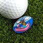 A close-up of a golf ball on green grass beside Logie Joe’s Golf’s "Douche Canoe" pin—an oval accessory with a cartoon dog paddling a pink canoe that adds humor and personality to your game.