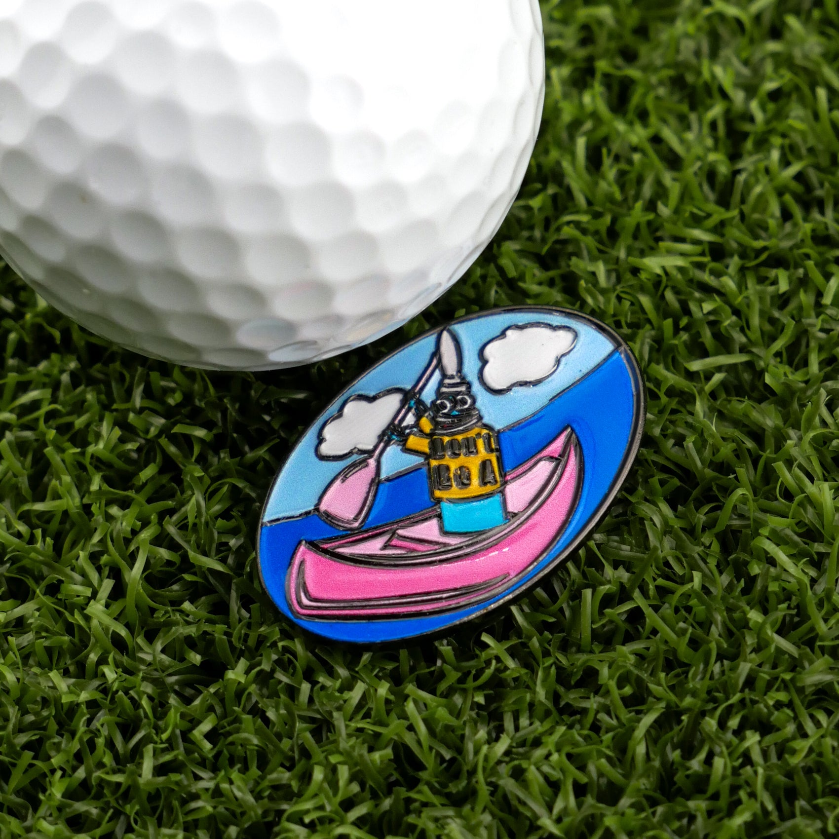 A close-up of a golf ball on green grass beside Logie Joe’s Golf’s "Douche Canoe" pin—an oval accessory with a cartoon dog paddling a pink canoe that adds humor and personality to your game.