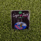 A Logie Joe's Golf Douche Canoe pin sits on artificial grass. Featuring a colorful cartoon design, it stands out among funny golf accessories and is displayed on a black square backing card for playful appeal.