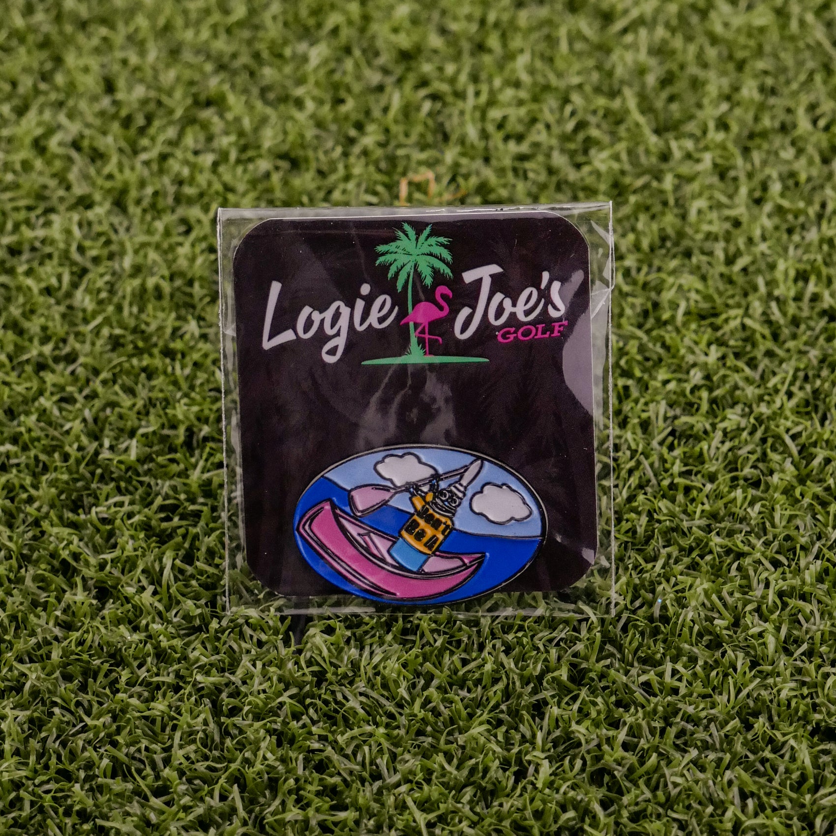 A Logie Joe's Golf Douche Canoe pin sits on artificial grass. Featuring a colorful cartoon design, it stands out among funny golf accessories and is displayed on a black square backing card for playful appeal.