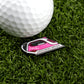 A close-up of a white golf ball on green grass beside a pink and black golf ball marker shaped like an iron, featuring Logie Joe's Golf Driving Iron—a unique premium golf accessory.