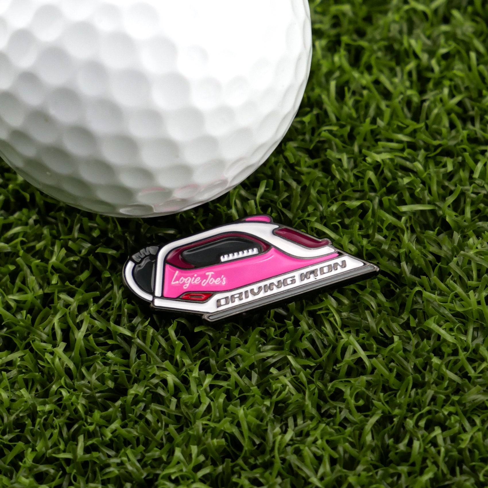 A close-up of a white golf ball on green grass beside a pink and black golf ball marker shaped like an iron, featuring Logie Joe's Golf Driving Iron—a unique premium golf accessory.