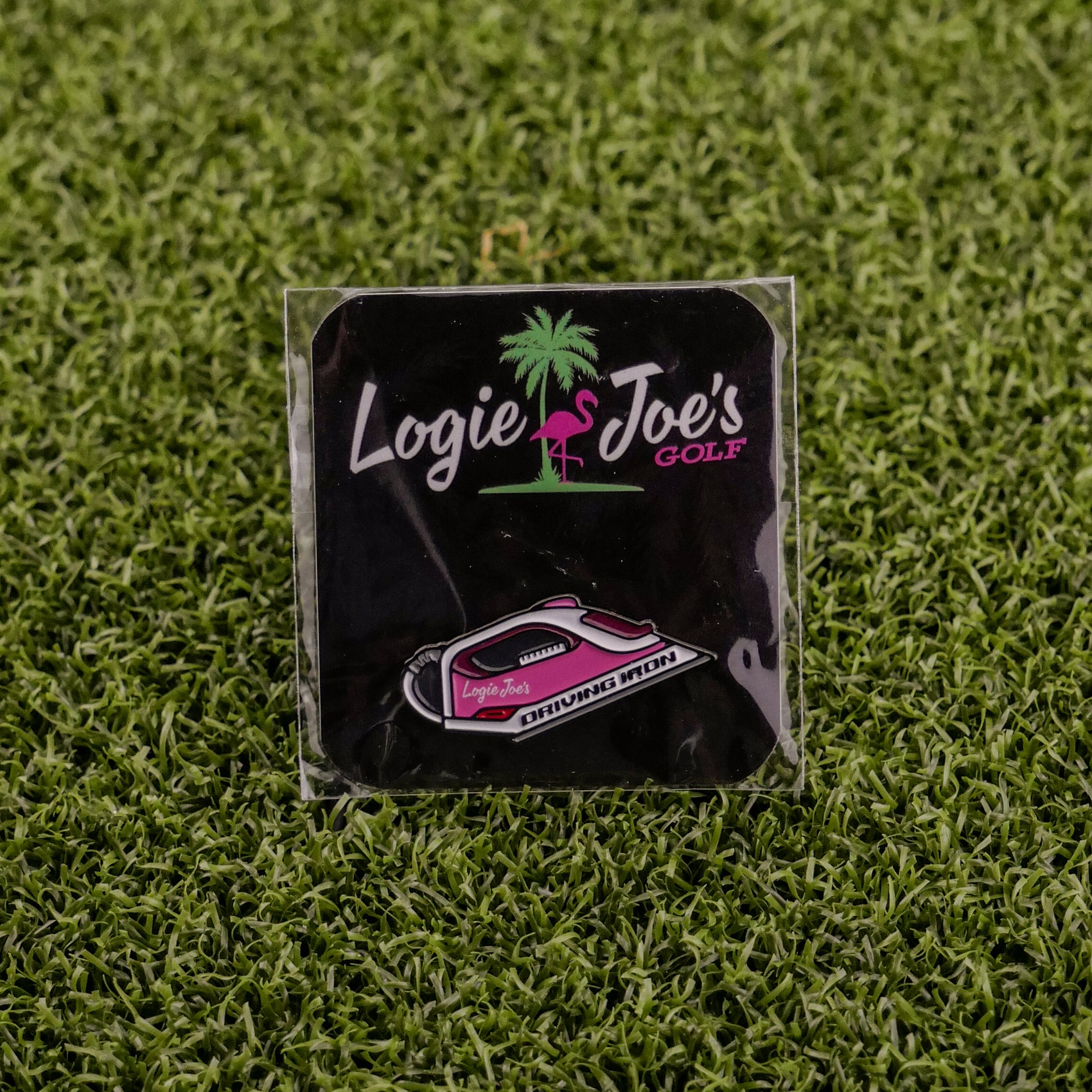 A Logie Joe's Golf Driving Iron pin displayed on artificial grass. This premium accessory features the brand logo in pink, green, and white—ideal for collectors or as a unique golf ball marker.